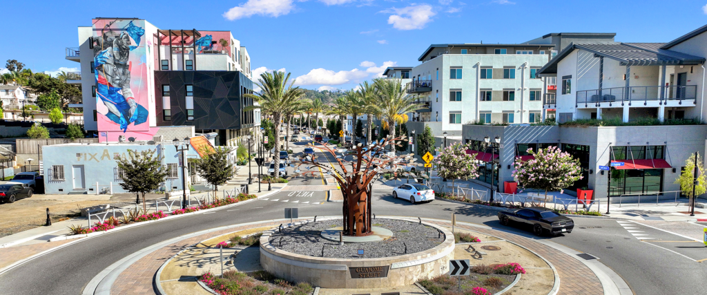 Roundabout with a rust-colored metal tree sculpture in the center, surrounded by palm trees and modern buildings.