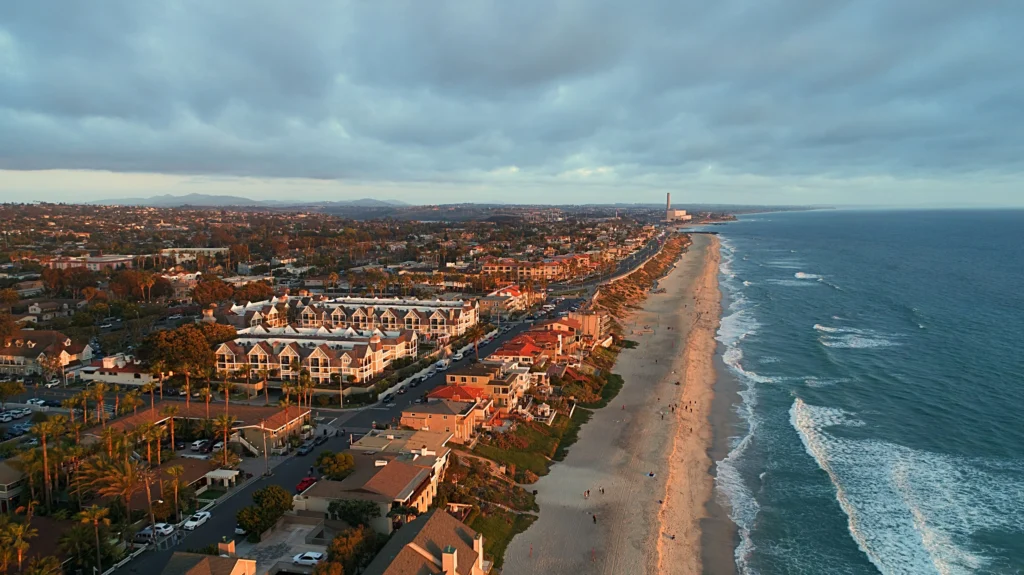 Aerial view of a coastal town along a long sandy beach with ocean on the right and a grid of houses and palm trees on the left at sunset/golden hour.