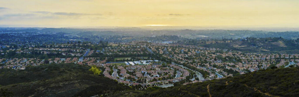 Aerial view of a suburban town set in rolling hills at sunset, with winding roads and red-roofed houses expanding to the horizon.