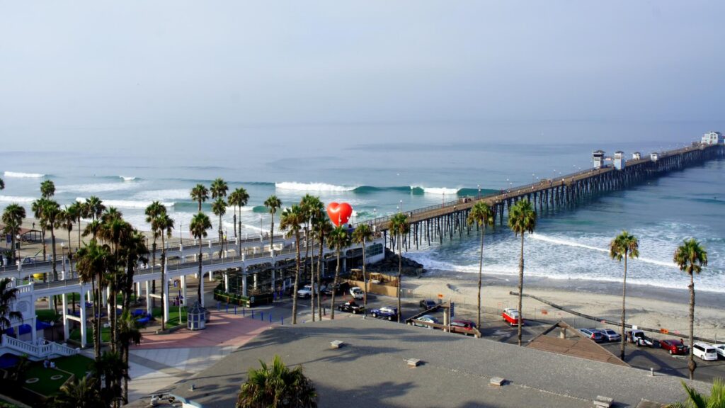 Pier extends into the ocean with palm trees along the beach and a red heart-shaped balloon near the center.